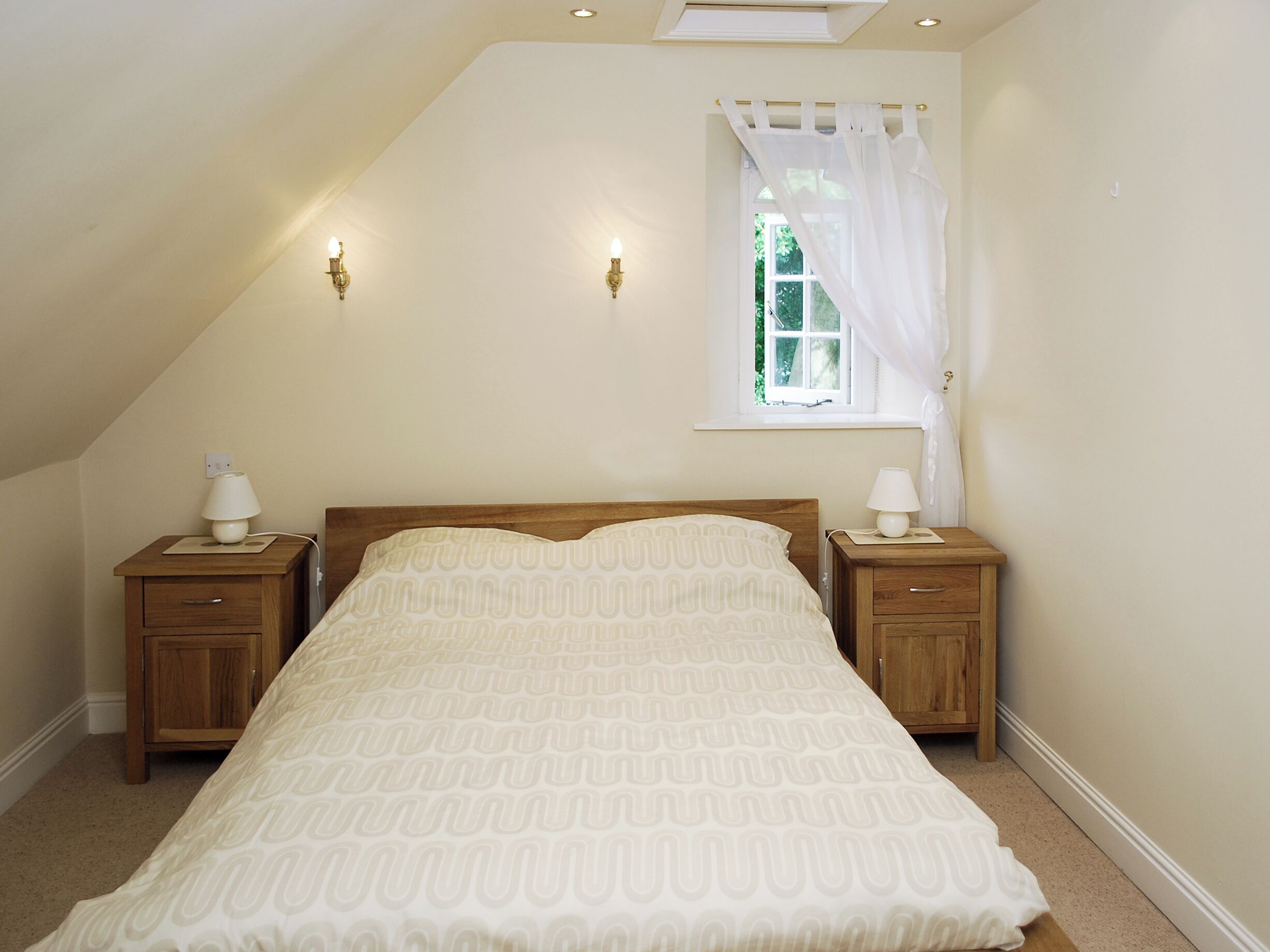 Une chambre avec un plafond en pente, un lit simple, deux tables de chevet avec des lampes, une fenêtre avec un rideau léger et une vue sur la verdure à l’extérieur.