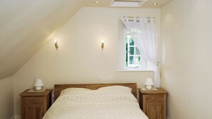 A bedroom with a sloped ceiling, a single bed, two bedside tables with lamps, a window with a sheer curtain, and a view of greenery outside.