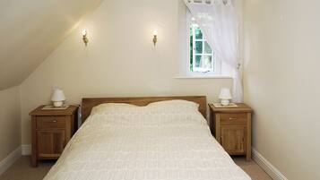 Une chambre avec un plafond en pente, un lit simple, deux tables de chevet avec des lampes, une fenêtre avec un rideau léger et une vue sur la verdure à l’extérieur.