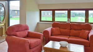 Ein Wohnzimmer mit einem roten Sessel und einer Couch, einem Holztisch und Blick auf einen Garten durch ein Fenster.