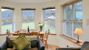 Una sala de estar acogedora con un sofá de cuero, una mesa de comedor de madera dispuesta para cuatro personas y ventanas amplias que ofrecen vistas a un campo y colinas.