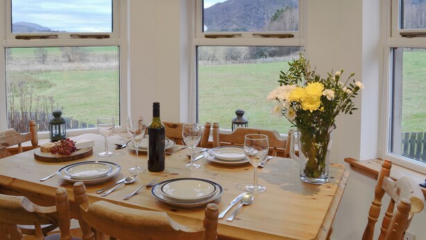 Una mesa de comedor dispuesta con platos, cubiertos y una botella de vino, con un florero y vistas a un campo verde a través de amplias ventanas.