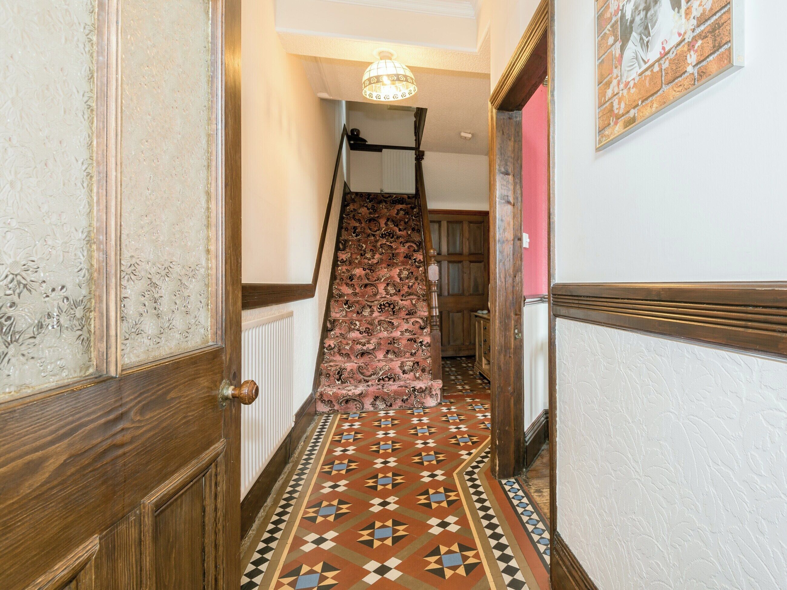 A hallway with a patterned floor, a wooden door, a staircase with a patterned carpet, and a hanging light fixture.
