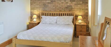 A bedroom with a wooden bed, two bedside tables with lamps, a brick wall, and a small wooden desk.