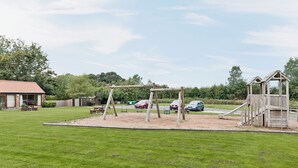 Un parque infantil con columpios y un tobogán, una mesa de picnic, y un edificio con techo rojo al fondo.