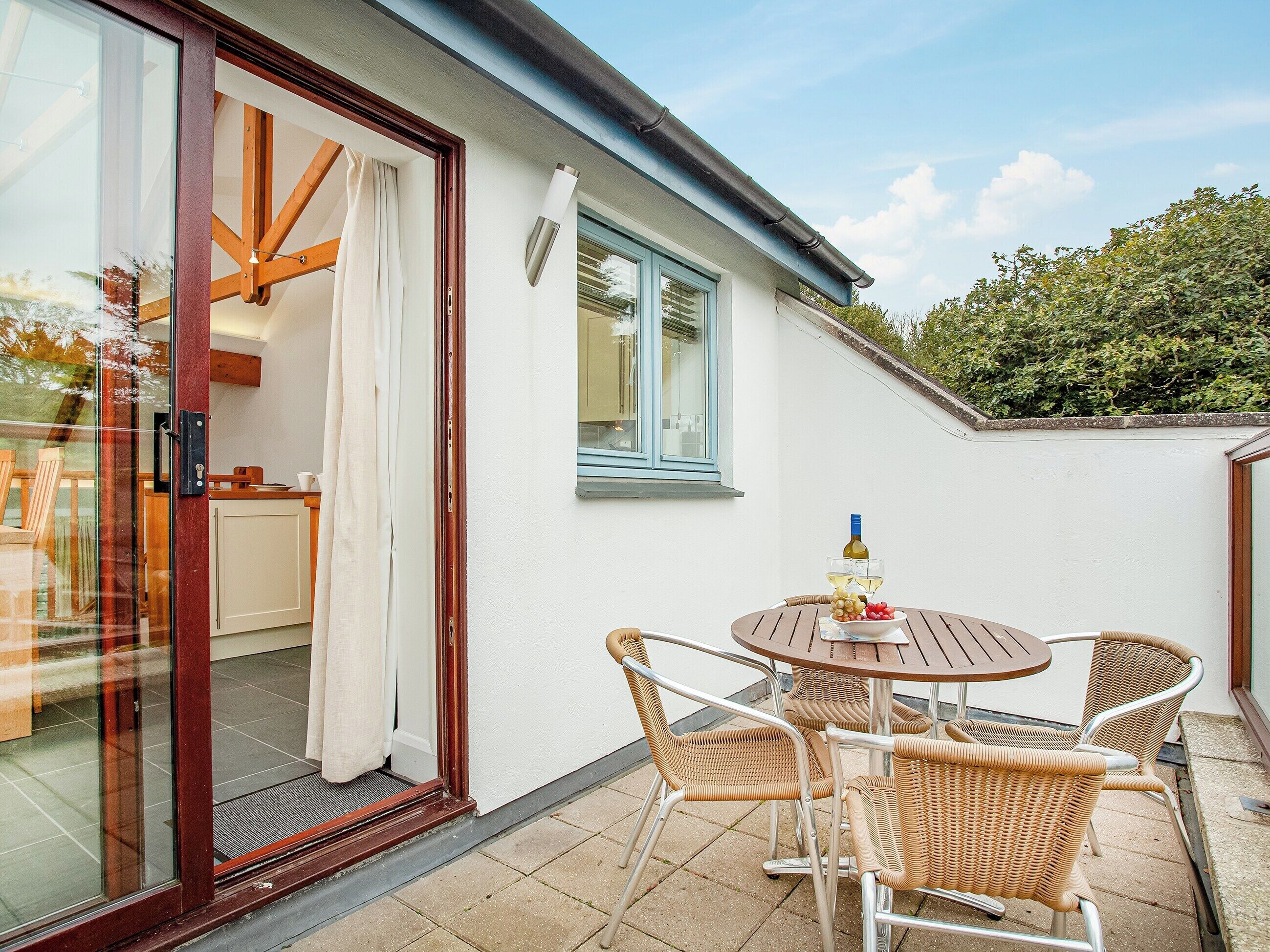 An outdoor patio with a round table and wicker chairs, a glass door leading to a room, and a window with blue shutters.
