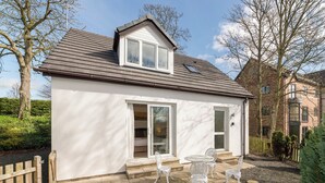 Ein kleines, eingeschossiges Haus mit weißer Fassade, geneigtem Dach und einer großen Schiebeglastür. Auf der Terrasse stehen drei weiße Gartenstühle und ein kleiner Tisch.