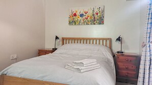 Un dormitorio con cama de madera, dos mesitas de noche con lámparas, una mesita auxiliar pequeña, un cuadro colgado en la pared y una cortina estampada.