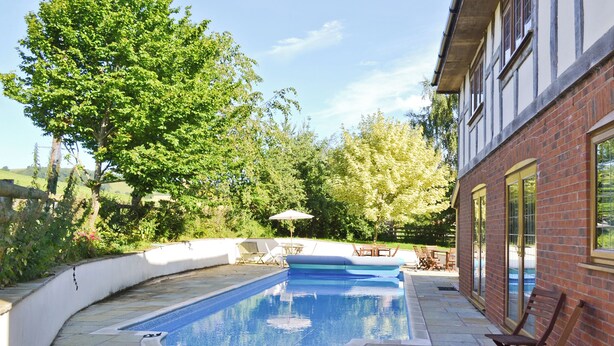 A swimming pool with a curved edge, surrounded by stone tiles, wooden chairs, and a white wall.