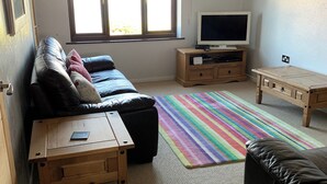 A living room with a sofa, a TV on a wooden stand, a small wooden table, and a colorful striped rug. - 3 bedroom accommodation in Benllech (Benllech)