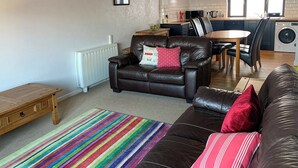 A living room with a sofa, a coffee table, and a striped rug. There is a dining area with a table and chairs, and a kitchen in the background. - 3 bedroom accommodation in Benllech (Benllech)
