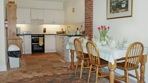 Une cuisine avec des meubles blancs, un pilier en briques et un coin repas avec des chaises en bois et une table dressée pour quatre personnes.