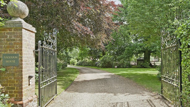 Une allée de gravier mène à une entrée fermée par un portail, sur lequel on peut lire l’inscription « Yaxham House ».