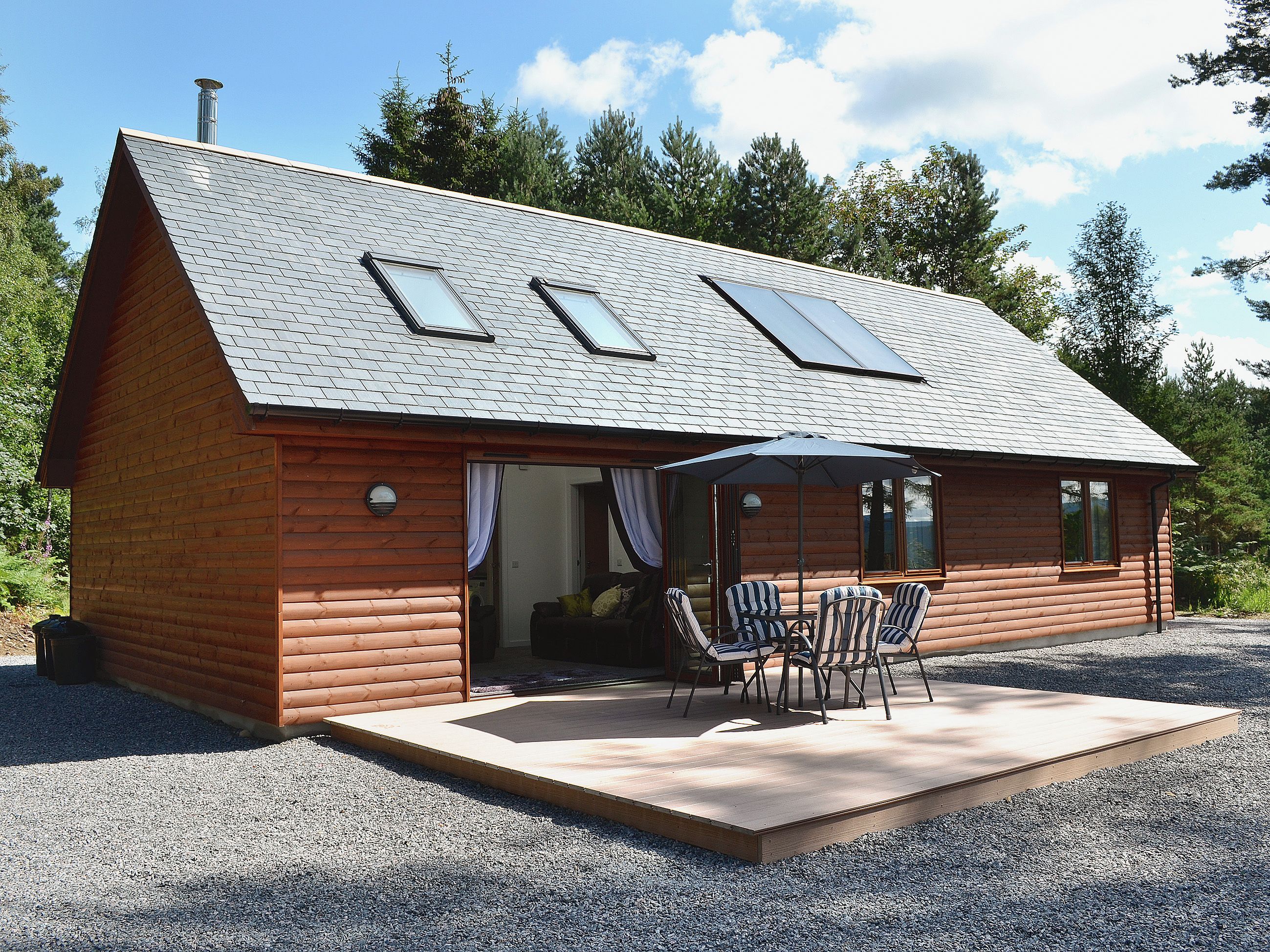 A log cabin with a sloped roof, skylights, a covered patio with chairs, and a gravel driveway.