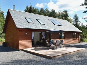 A log cabin with a sloped roof, skylights, a covered patio with chairs, and a gravel driveway.