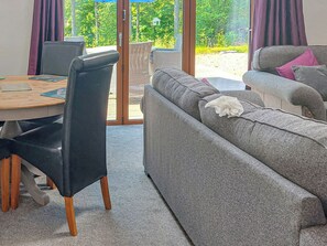 A room with a grey sofa, a dining table with chairs, and a view through a glass door to an outdoor area with greenery.