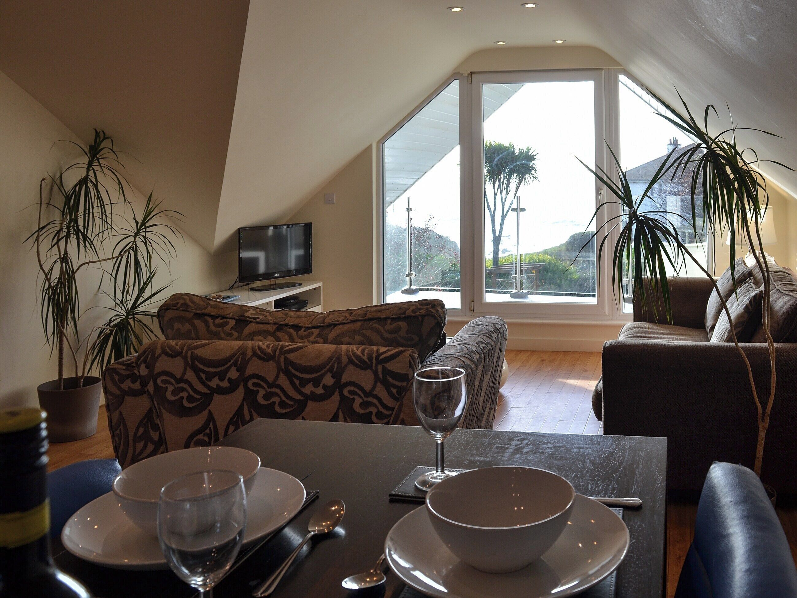 A dining area with a table set for two, a television on a stand, and large windows offering a view of the outdoors.