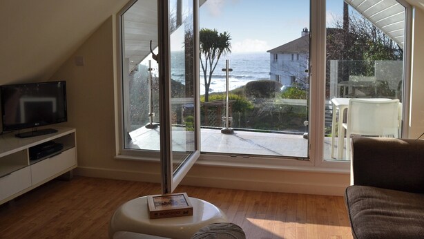 A room with a large bay window overlooking the ocean, a television on a white stand, and a small table with a board game. - The lounge is a room with a view. (Heybrook Bay, near Plymouth)