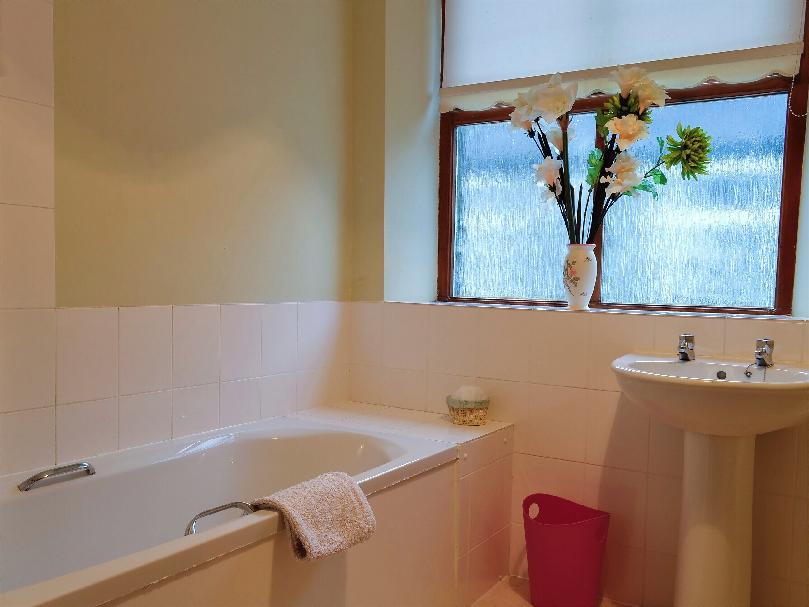 A bathroom with a bathtub, a sink, and a window with a vase of flowers.
