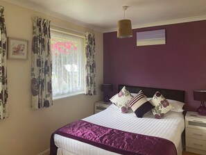 A bedroom with a bed, a nightstand, a lamp, a window with curtains, and a painting on the wall.