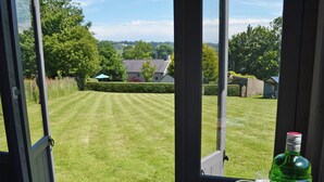 A view through a window of a well-maintained lawn, a table with a book and a bottle, and a glass of water. - 3 bedroom accommodation in Llangoed, near Beaumaris (Llangoed, near Beaumaris)