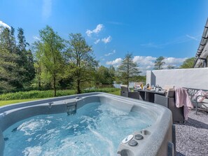 A hot tub with a table and chairs, surrounded by greenery and trees, with a clear sky in the background.