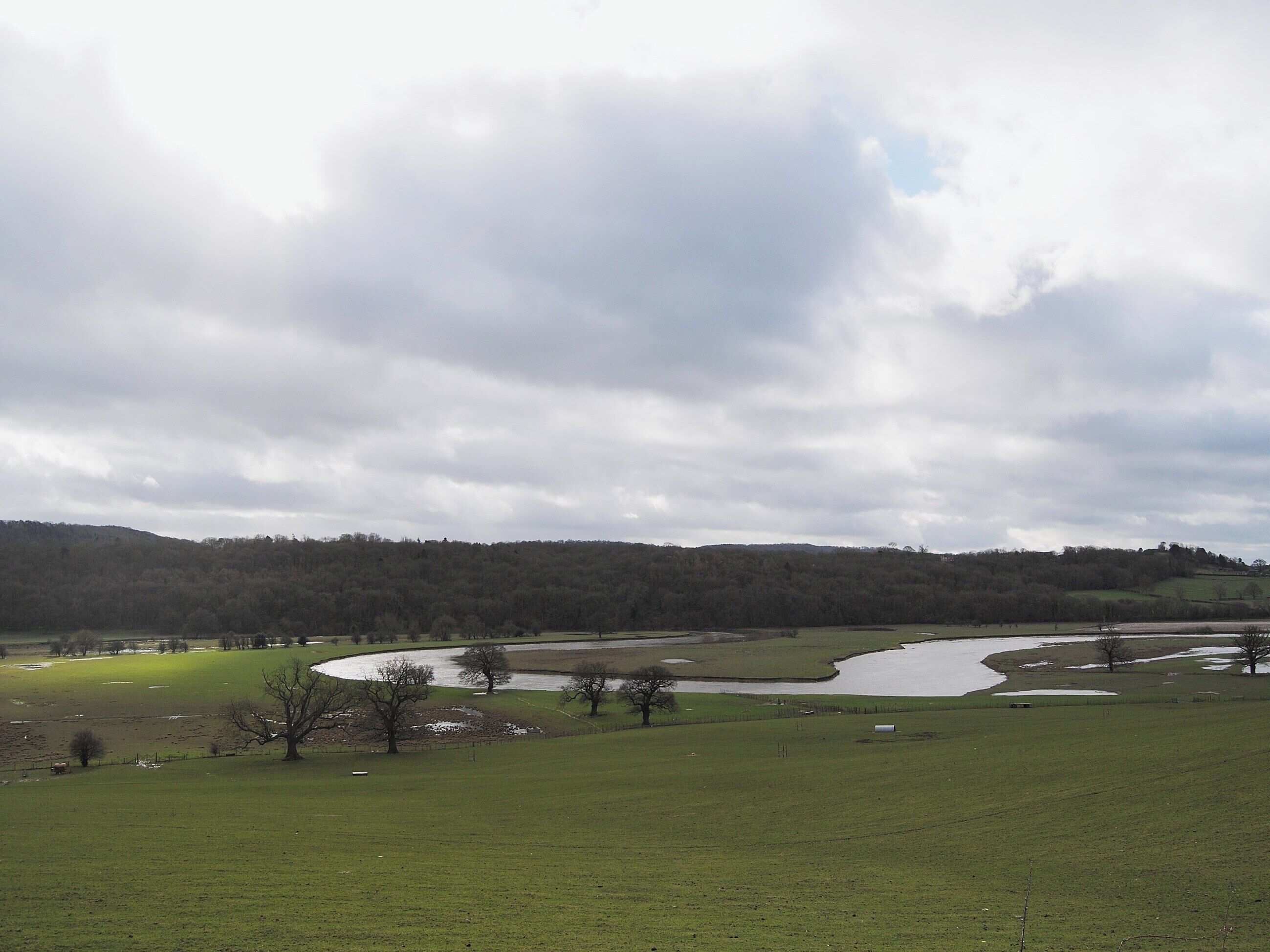 Een landelijk landschap met een rivier, open velden en een bebost heuvel op de achtergrond.