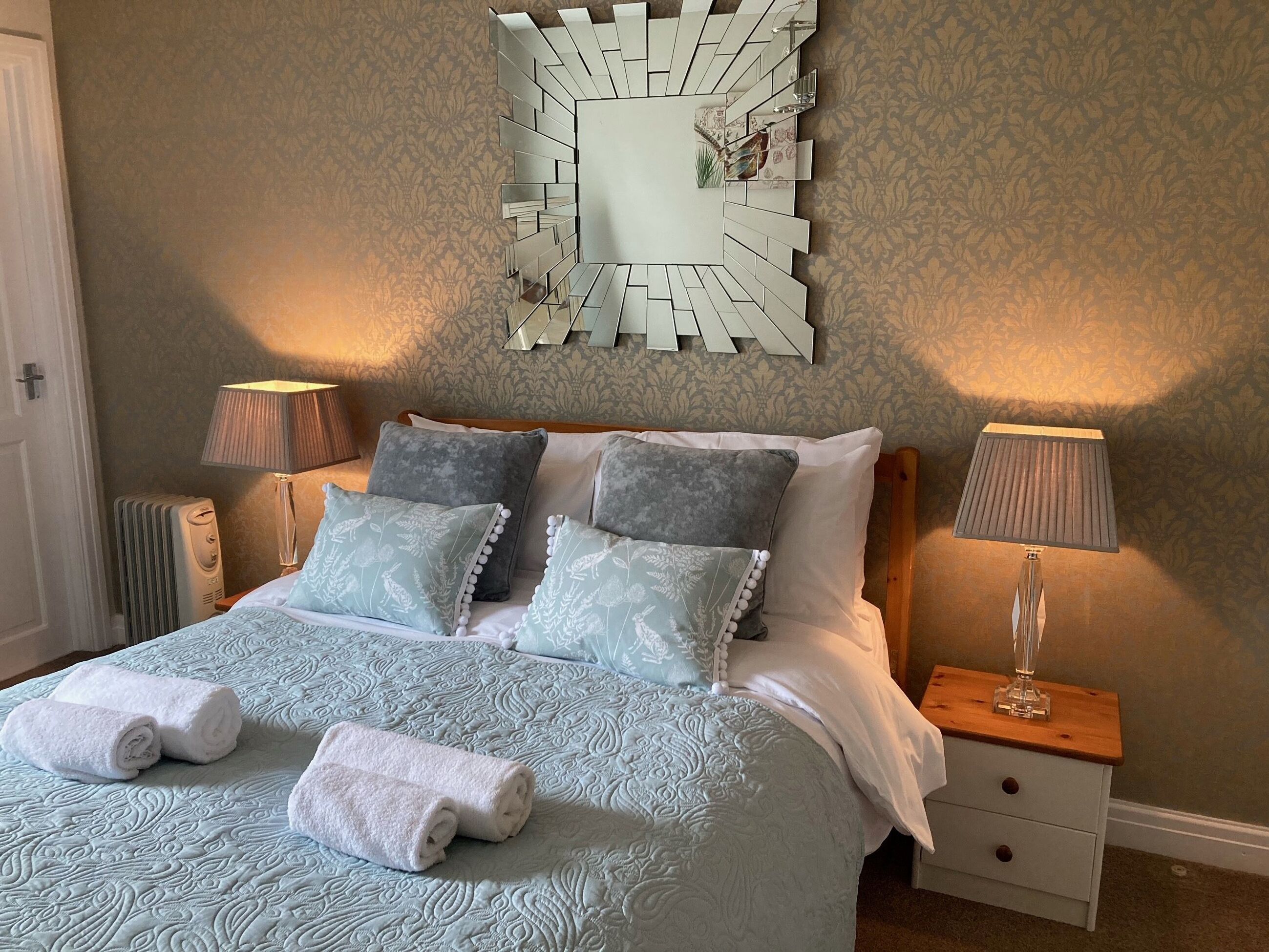 Un lit bien fait, avec une literie à motifs, deux lampes de chevet, un miroir décoratif et un radiateur.