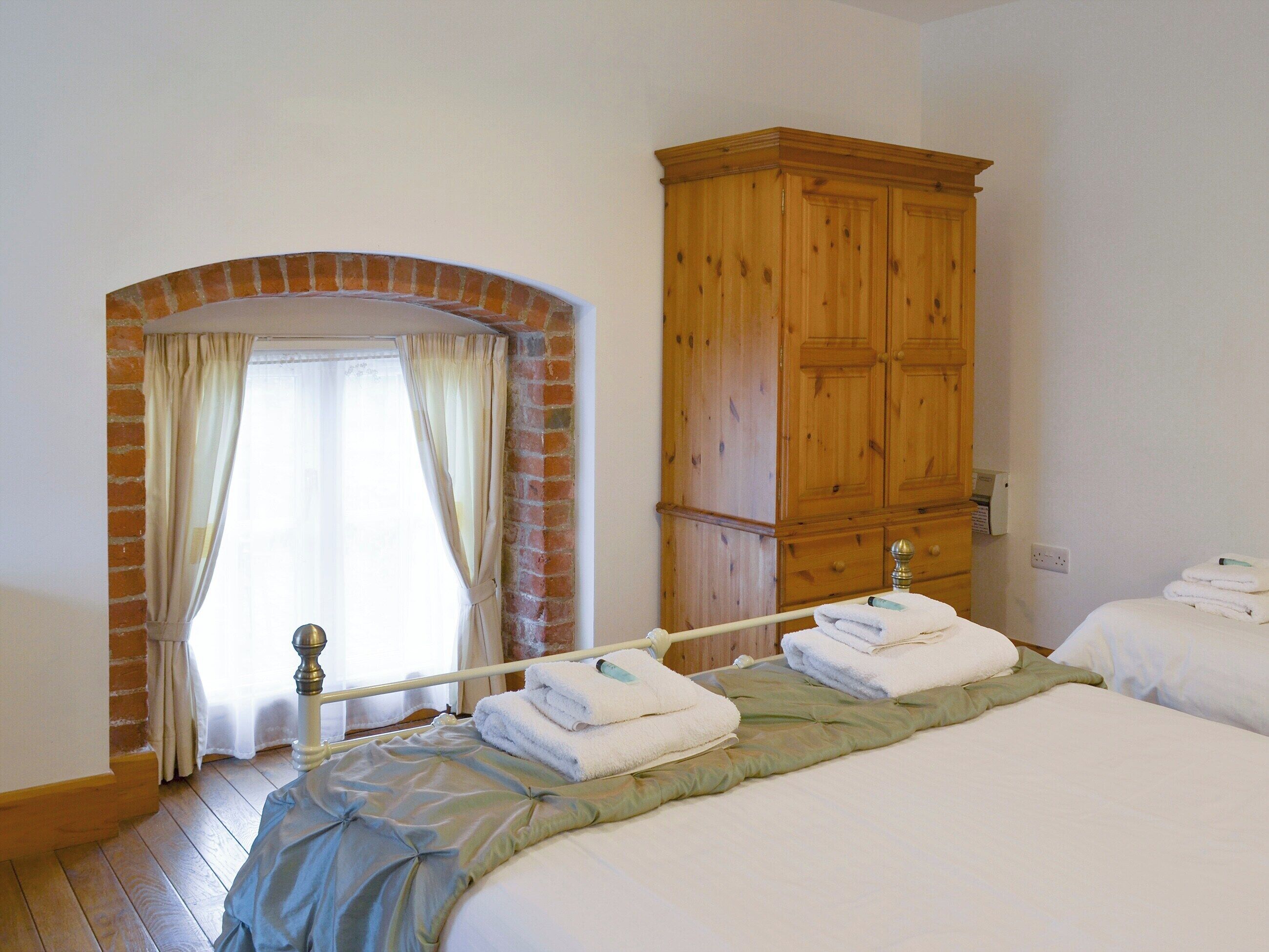 A bedroom with two beds, a wooden wardrobe, a brick archway, and a window with curtains.