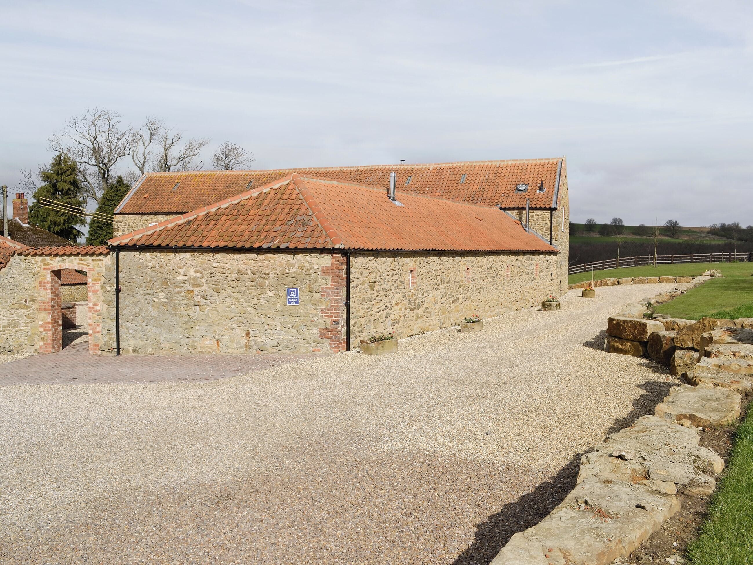 A stone building with a tiled roof, a gravel driveway, and a stone wall with a wooden fence in the background.