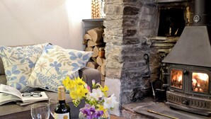 Ein gemütliches Wohnzimmer mit Kamin aus Stein, einer Couch mit gemusterten Kissen, einem Tisch mit Weinflasche, Gläsern und Obst.