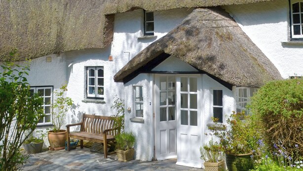Ein traditionelles weißes Cottage mit Strohdach, einer hölzernen Bank und Topfpflanzen.