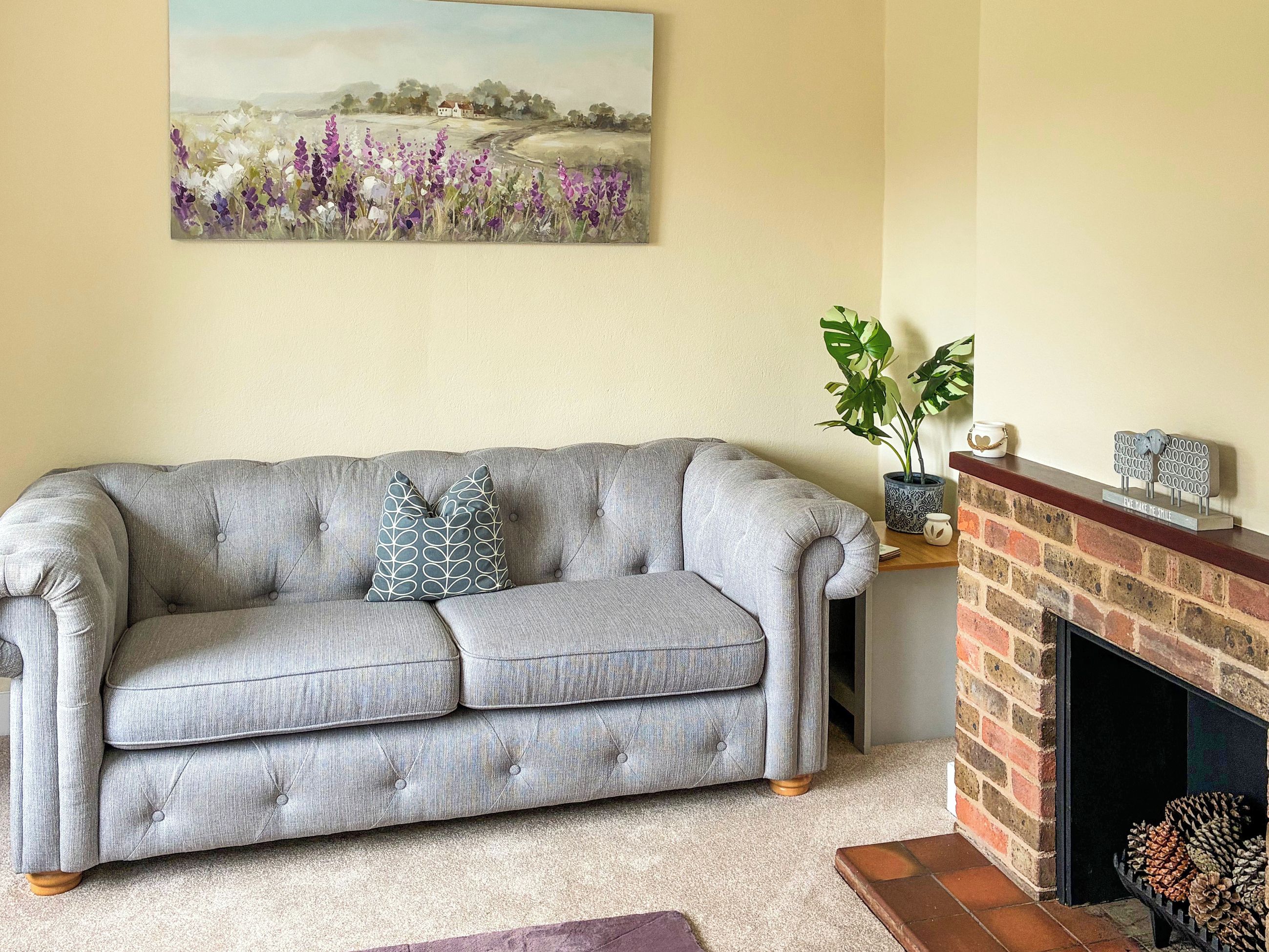A grey upholstered sofa with two patterned cushions, a painting of a landscape, a potted plant, and a brick fireplace with pine cones.