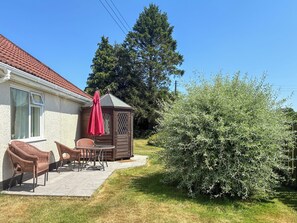 Une maison avec un patio, un abri de jardin en bois et un grand buisson.