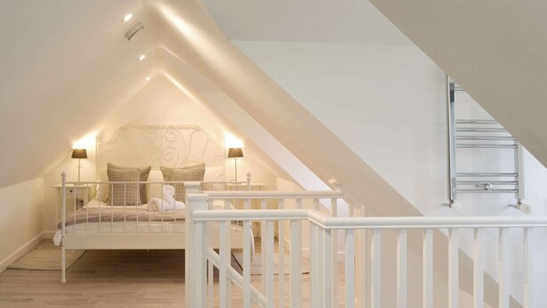 A bedroom with a sloped ceiling, a bed with white bedding, two bedside lamps, and a white railing. - 3 bedroom accommodation in Helstone, near Camelford (Helstone, near Camelford)
