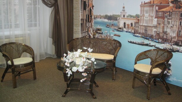Une pièce décorée d’une grande fresque représentant Venise, avec deux fauteuils en osier, une petite table sur laquelle est posé un vase de fleurs et une fenêtre agrémentée de rideaux.