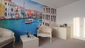 Une pièce décorée d’une fresque représentant le Grand Canal de Venise, avec un canapé blanc, une petite table sur laquelle se trouvent une bouteille et des verres, et un coin cuisine.