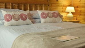 Una cama bien hecha con cubrecama blanco y almohadas decorativas, cabecera de madera, mesita de noche con lámpara y una nota sobre la cama.