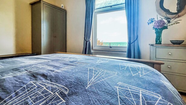 Une chambre à coucher avec un lit, une armoire, une fenêtre avec des rideaux et un vase rempli de fleurs posé sur une commode.