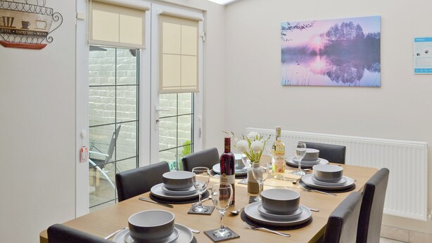 Un coin repas avec une table dressée pour six personnes, comprenant des assiettes, des bols, des verres et des couverts. Des chaises sont disposées autour de la table, et une fenêtre avec des stores est visible.