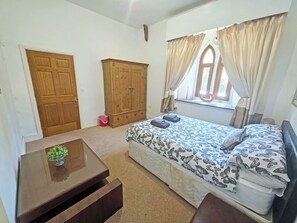 A bedroom with a bed, a wooden wardrobe, a small table with a plant, and a window with curtains. - St Albans Church - 28165 (Treherbert, near Treorchy)