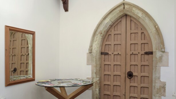 A room with a round glass-top table, a wooden door with a stone arch, and a mirror on the wall.