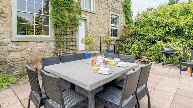 A stone building with ivy, a table set for breakfast, and a barbecue grill.
