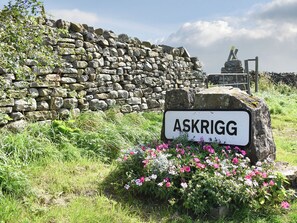Un muro de piedra con un letrero que dice “ASKRIGG”, rodeado de hierba y flores.