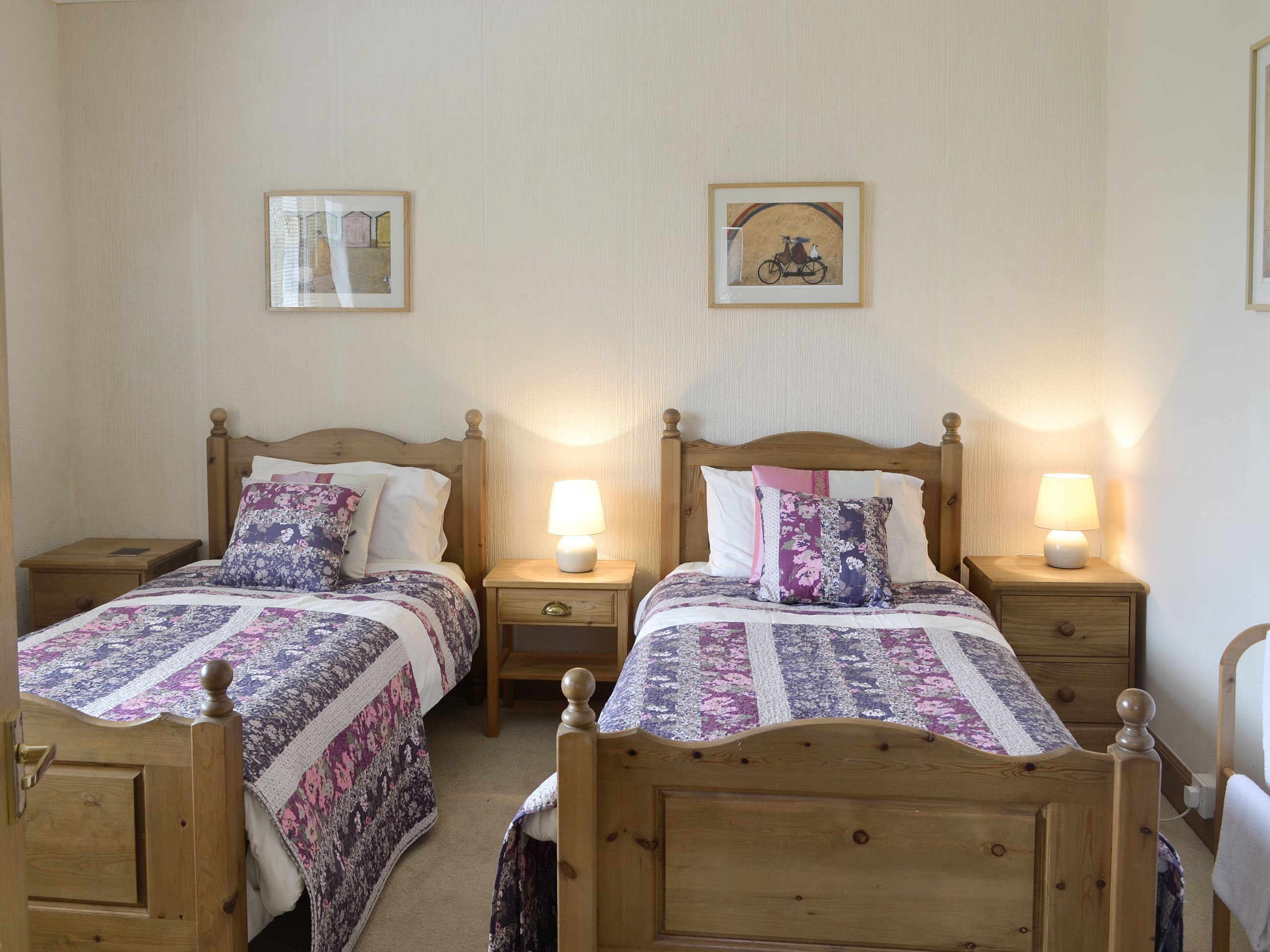 A double room with two wooden beds, each with a nightstand and a lamp. There are framed pictures on the wall above the beds.