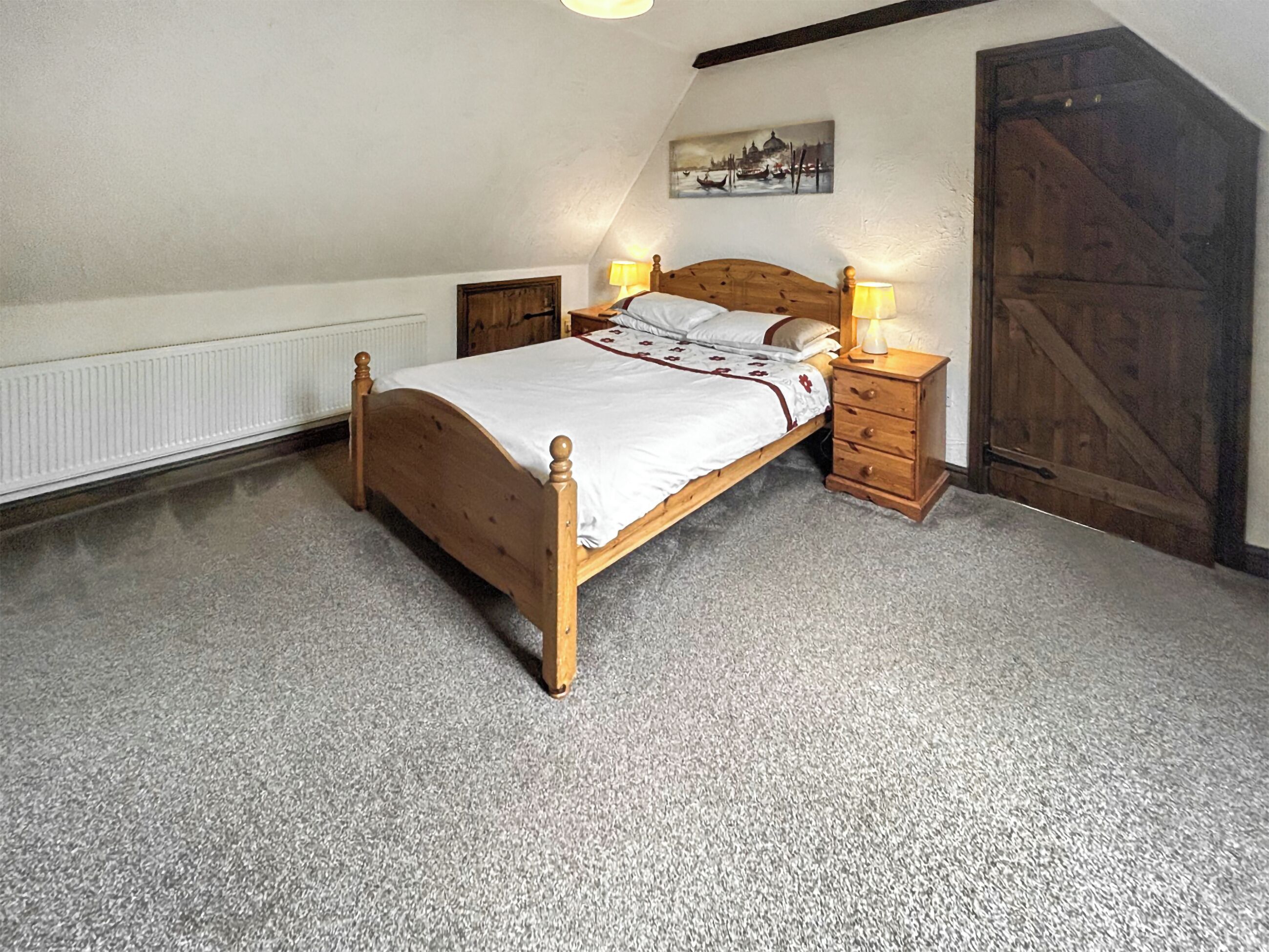Une chambre avec un lit en bois, deux tables de chevet avec des lampes, un radiateur, une porte en bois et un tableau accroché au mur.