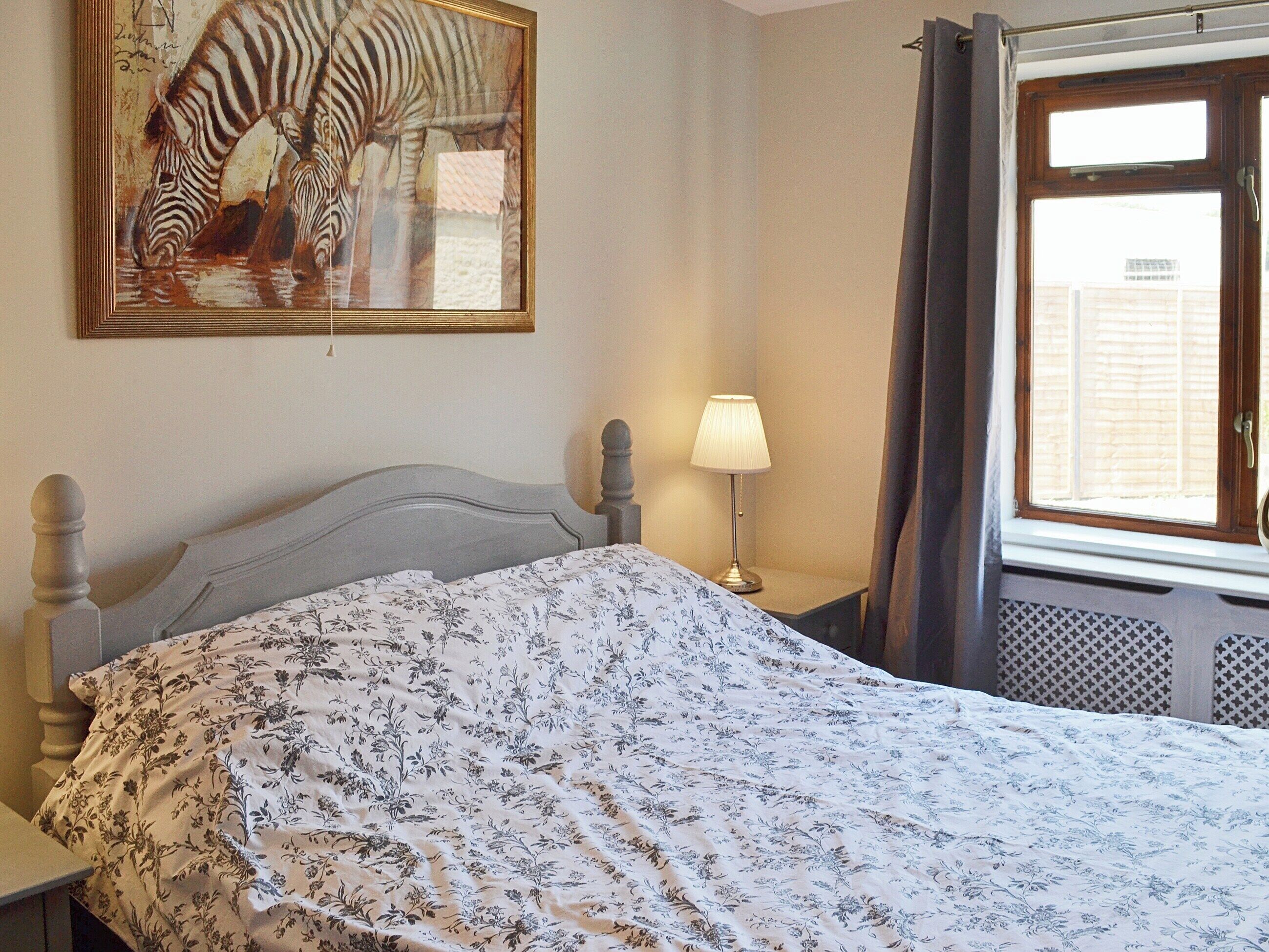 A bedroom with a bed, a nightstand, a lamp, a window with curtains, and a zebra painting on the wall.