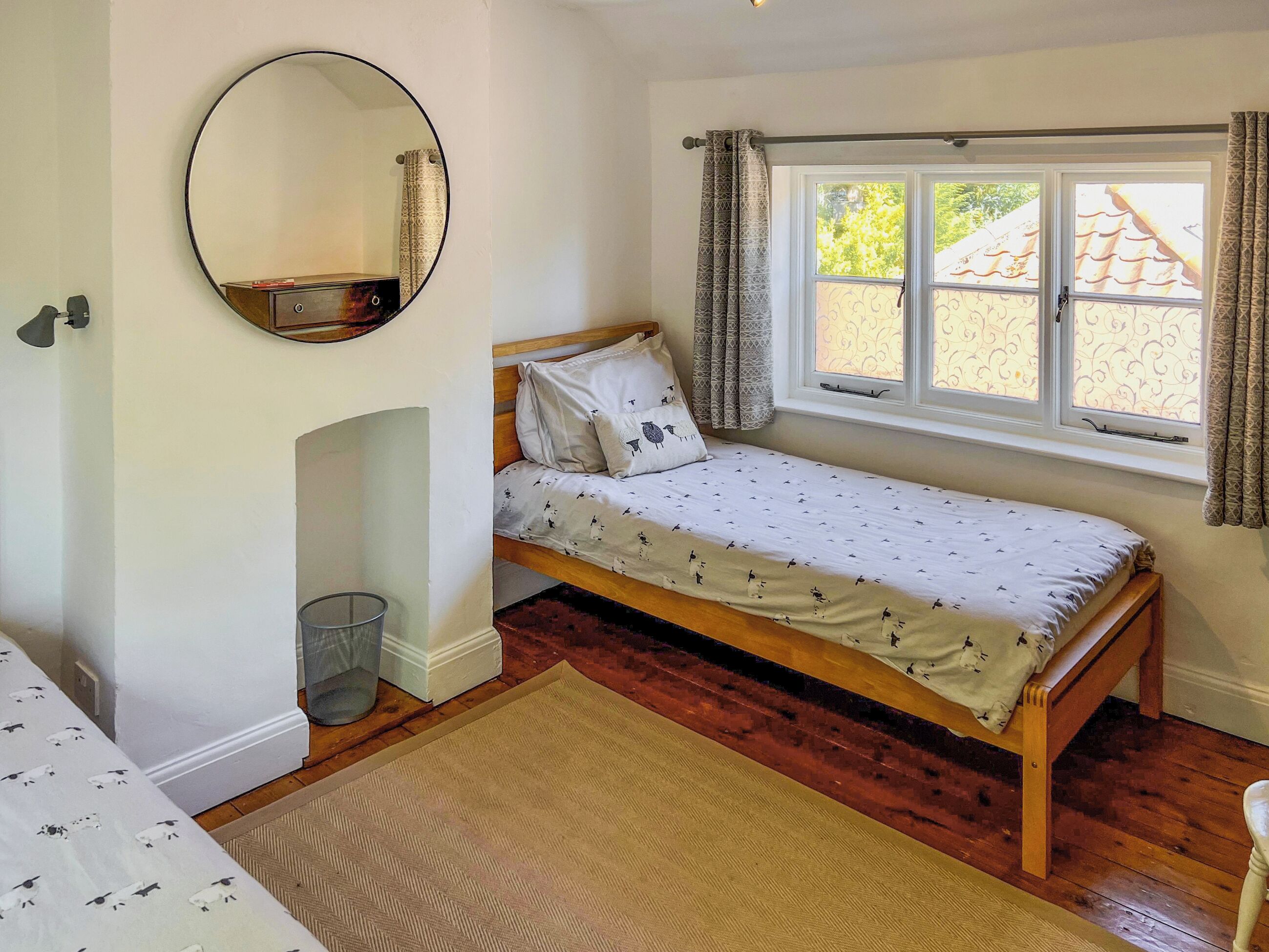 Un dormitorio con una cama individual, una ventana pequeña con cortinas, un espejo redondo y suelo de madera.