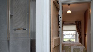 Ein Badezimmer mit Duschabtrennung aus Glas, einer Holztür, die in einen Schlafbereich mit einem blumengedeckten Bett führt, und einem Fenster mit Vorhängen.