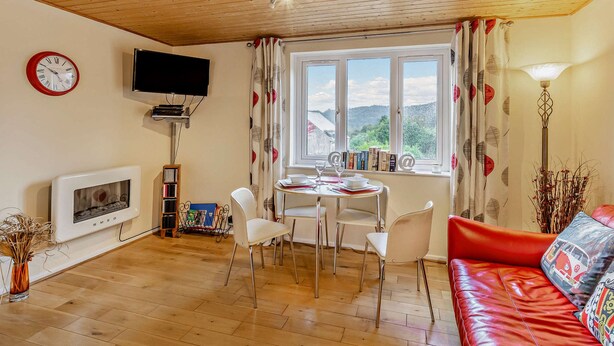 A cozy living room with a wooden ceiling, a fireplace, a TV, a dining area with chairs, a red sofa, and a window with a view of the hills.